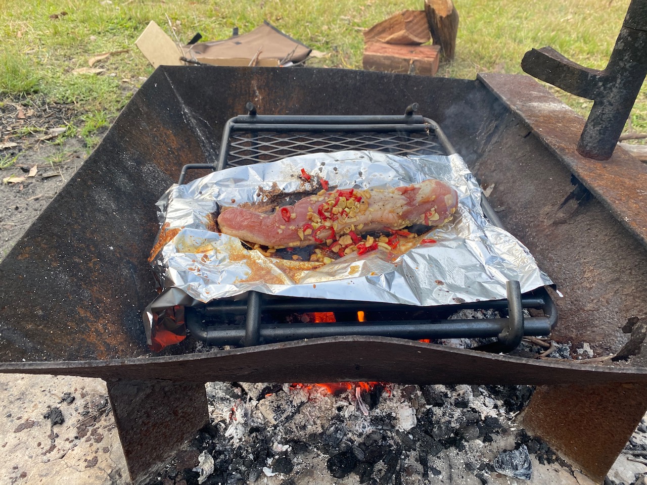 A camping grill over a wood fire, with a large piece of pork cooking on a sheet of aluminium foil