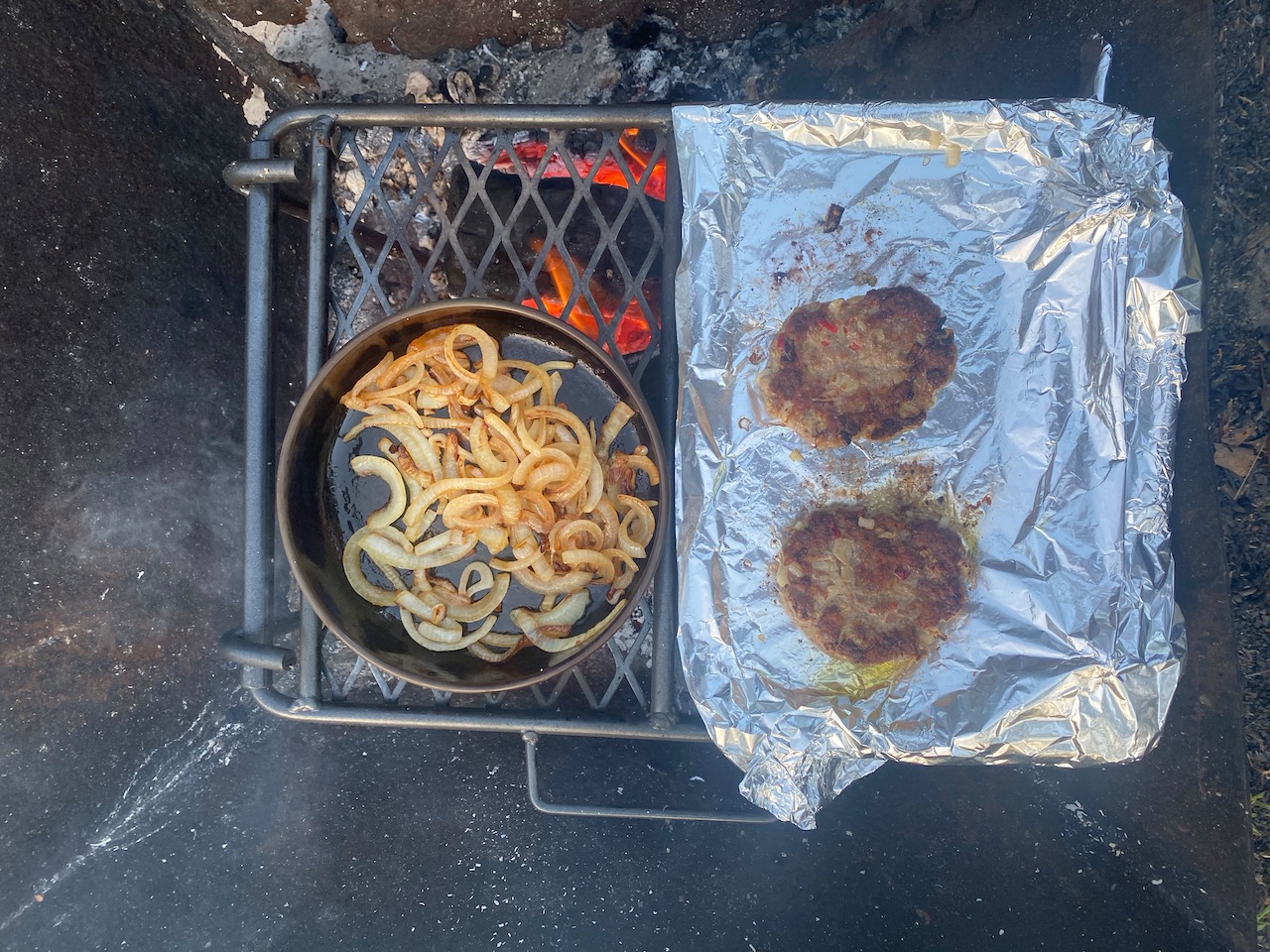 A camping grill over a wood fire, with two hamburger patties and a circular pan with fried onions