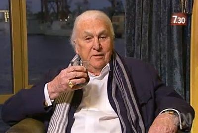 John Laws, with a glass of something, in a chair, wearing a scarf and a big ring