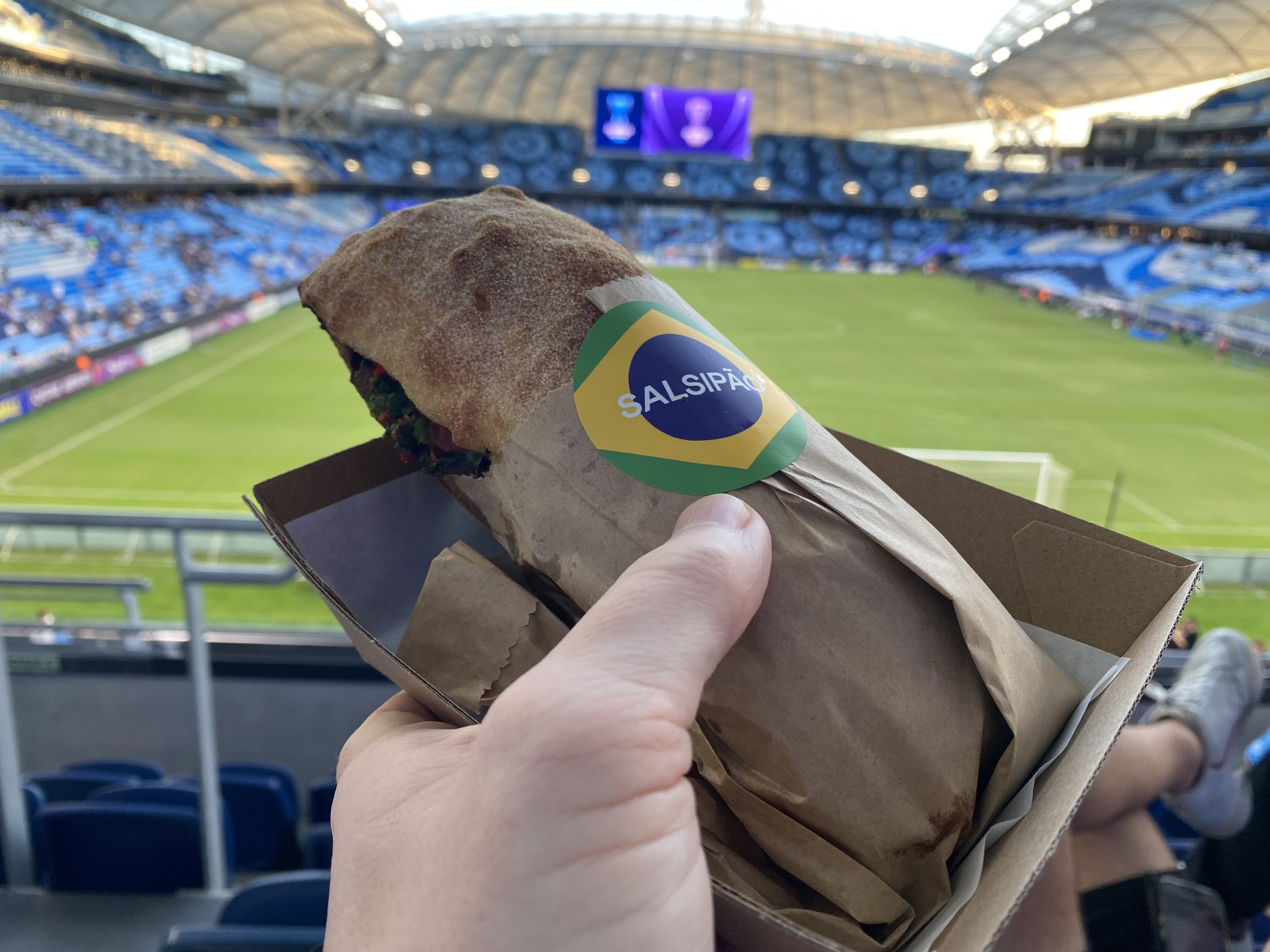 My hand holding a pork roll in a paper bag and a box, marked ' Salsipão' with a Brazilian flag, with a football stadium out of focus in the background