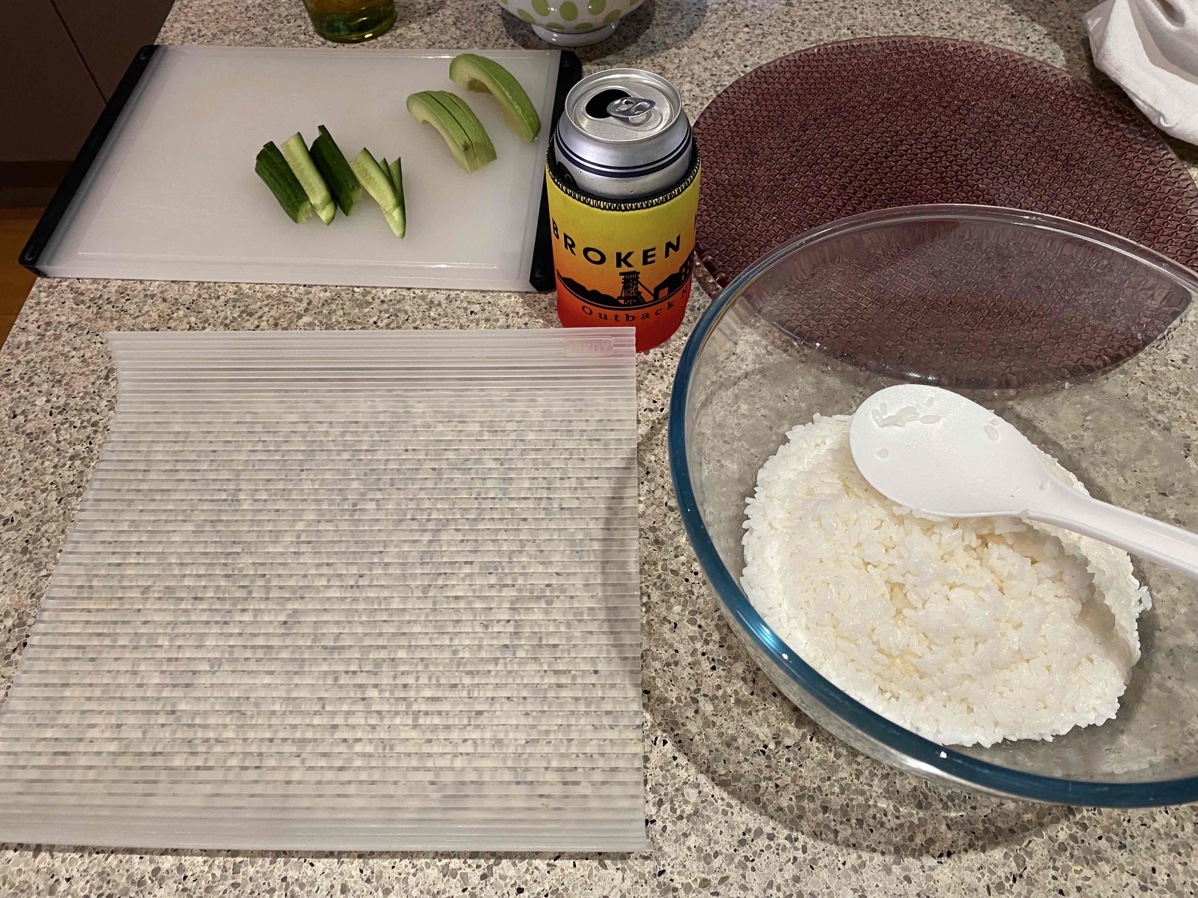 Sushi rice, a roller, some chopped avocadoes and cucumbers, and a can of beer